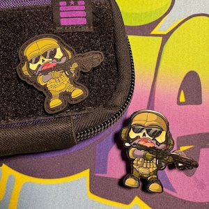 May include: Two cartoon character patches, one on a black zippered pouch and one on a blue and yellow background. Both patches are of a cartoon character wearing a helmet, sunglasses, and a camouflage uniform, holding a rifle. The character has a skull face with a pink tongue sticking out.