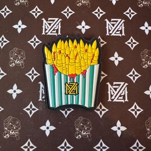 May include: A green and white striped container with a red drip, filled with yellow bullet-shaped fries. The container has a black logo with the letters "MZ" inside.