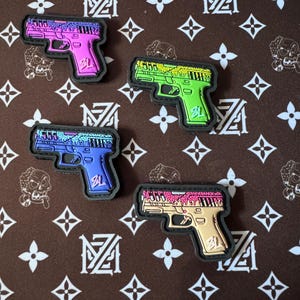 Puede incluir: Cuatro parches de pistola de diferentes colores con un diseño de goteo. Los parches son negros, rosas, verdes y morados. Cada parche tiene las letras "SL" en la parte inferior.