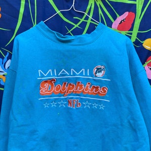 Könnte beinhalten: Ein türkisblauer Pullover mit den Worten "Miami Dolphins" und "NFL", die in Orange und Weiß gestickt sind.