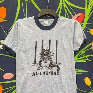 May include: A light blue t-shirt with a navy blue ringer neck. The shirt features a black and white illustration of a cat in a jail cell with the text "AL CAT RAZ" underneath.