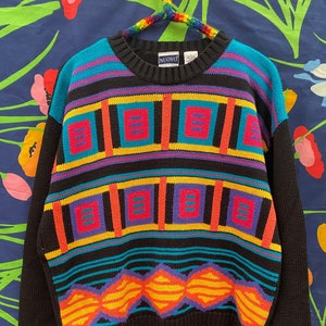 May include: Black sweater with a colorful geometric pattern. The pattern features squares, diamonds, and stripes in shades of blue, red, yellow, orange, and purple. The sweater has a crew neck and long sleeves. The brand name "NUOVO" is visible on the tag.