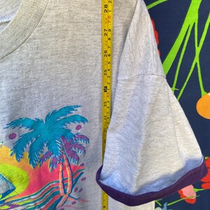 May include: A gray t-shirt with a colorful graphic of a palm tree and a sunset. The shirt has a purple ribbed trim around the neck and sleeves. The shirt is measured with a tape measure, showing the length of the sleeve.