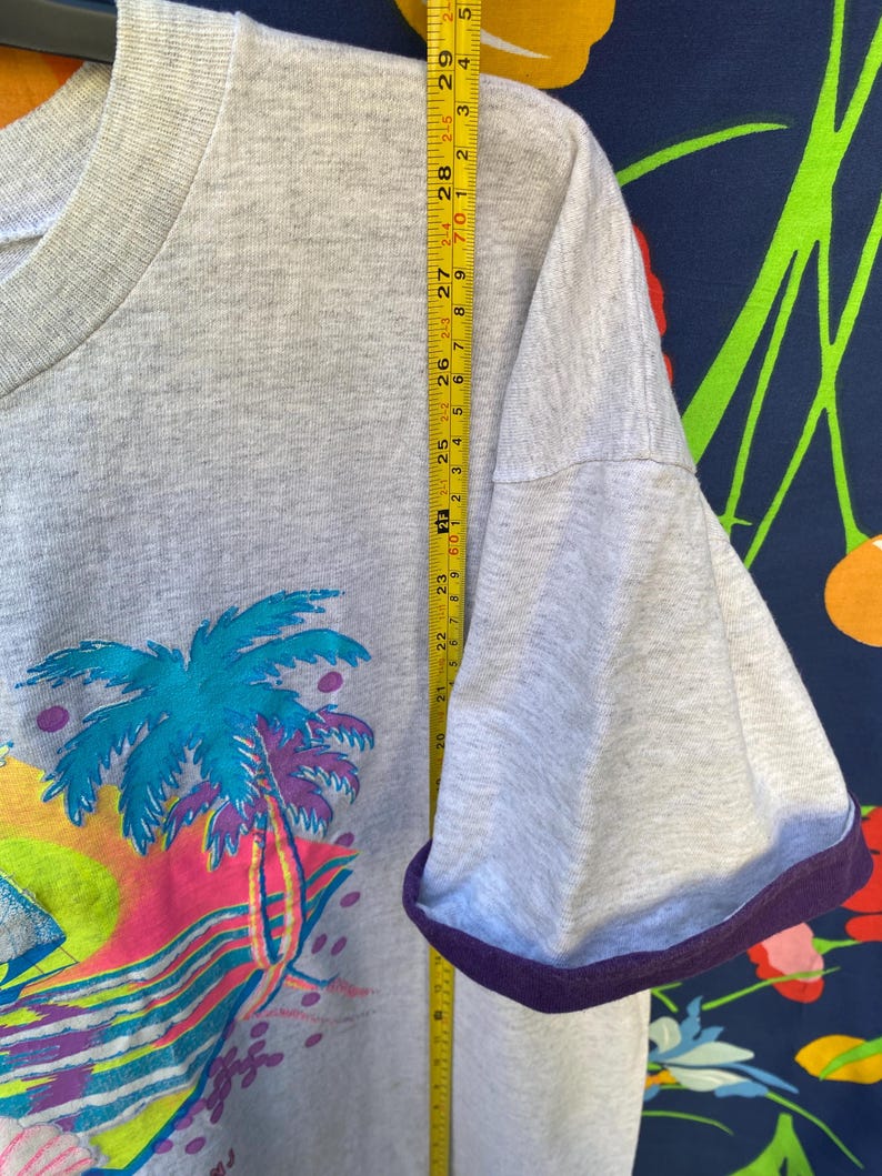 May include: A gray t-shirt with a colorful graphic of a palm tree and a sunset. The shirt has a purple ribbed trim around the neck and sleeves. The shirt is measured with a tape measure, showing the length of the sleeve.