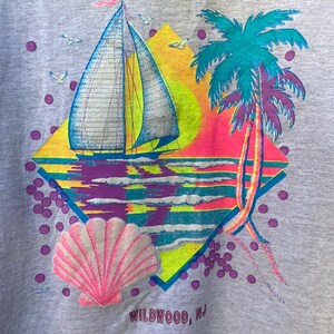 May include: A grey t-shirt with a colorful graphic design of a sailboat, palm trees, a seashell, and a sunset. The text "WILDWOOD, NJ" is printed on the bottom of the design.
