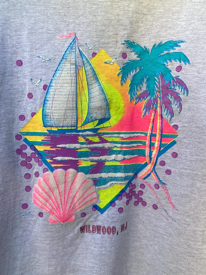 May include: A grey t-shirt with a colorful graphic design of a sailboat, palm trees, a seashell, and a sunset. The text "WILDWOOD, NJ" is printed on the bottom of the design.