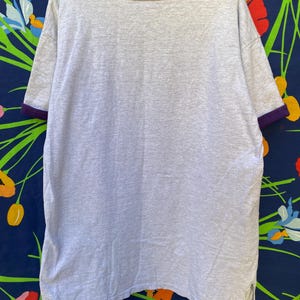 May include: A gray t-shirt with purple trim around the neck and sleeves. The shirt is made of a soft, comfortable material.