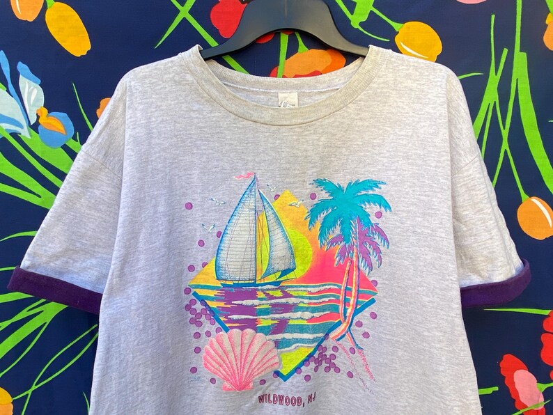 May include: Gray t-shirt with a colorful graphic of a sailboat, palm tree, and seashell. The graphic is in a diamond shape with the text "Wildwood, NJ" at the bottom.