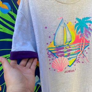 May include: A light gray t-shirt with a colorful graphic print of a sailboat, palm trees, and a seashell. The graphic also includes the text "WILDWOOD, NJ". The shirt has purple trim on the sleeves.