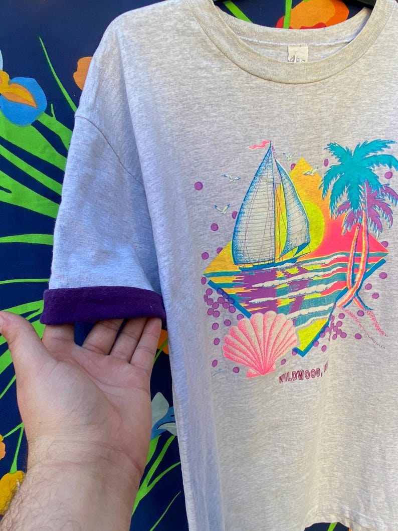 May include: A light gray t-shirt with a colorful graphic print of a sailboat, palm trees, and a seashell. The graphic also includes the text "WILDWOOD, NJ". The shirt has purple trim on the sleeves.