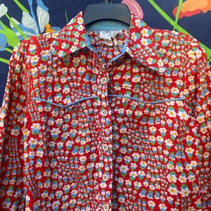Pode incluir: Camisa vermelha com um pequeno padrão floral em branco, azul e amarelo. Tem gola pontiaguda e debrum azul claro. Abotoada e pendurada num cabide preto. Feita de tecido leve.