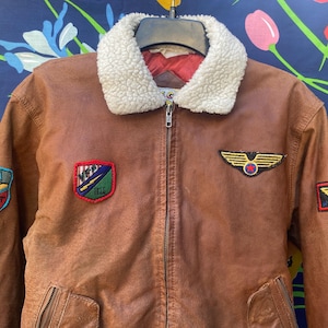 May include: A brown leather bomber jacket with a faux fur collar. The jacket has several patches, including a "USA Liberty" patch, a "V.F.P. Test Team" patch, and two patches with wings.