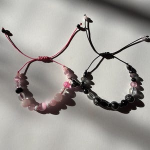 May include: Two beaded bracelets. One bracelet features pink and clear beads with a pink cord. The other bracelet has black and clear beads with a black cord. Both bracelets have adjustable closures.