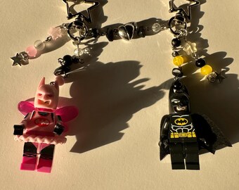 May include: Two keychains featuring Lego-style figures. One is a pink and black figure with a pink skirt and wings. The other is a black Batman figure with a yellow emblem. Both have star-shaped clasps and beaded accents.