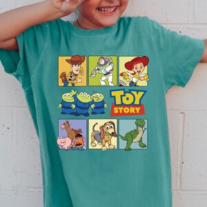 May include: Teal t-shirt featuring a colorful graphic of characters from the animated film "Toy Story." The design includes images of Woody, Buzz Lightyear, Jessie, the Aliens, Hamm, Slinky Dog, and Rex, with the "Toy Story" logo.