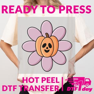 May include: A light-coloured t-shirt with a large cartoon design. The design features a smiling orange pumpkin with a jack-o'-lantern face at the centre of a pink flower. The words "READY TO PRESS" and "HOT PEEL DTF TRANSFER" are printed on the shirt.