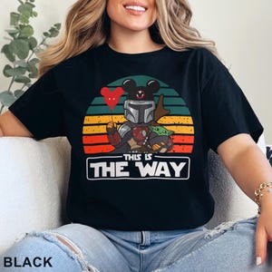 May include: Black t-shirt featuring a graphic of a Mandalorian character holding a red Mickey Mouse balloon and Grogu, set against a sunset design. The text "THIS IS THE WAY" is displayed in a white box. The word "BLACK" is at the bottom.