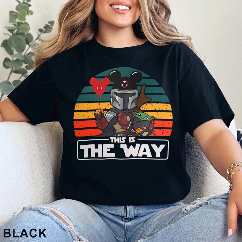 May include: Black t-shirt featuring a graphic of a Mandalorian character holding a red Mickey Mouse balloon and Grogu, set against a sunset design. The text "THIS IS THE WAY" is displayed in a white box. The word "BLACK" is at the bottom.
