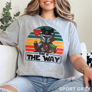 May include: Sport grey t-shirt featuring a graphic of a Mandalorian-inspired character with a Mickey Mouse head, holding a heart-shaped balloon, and Grogu. The design is set against a retro sunset with the text "THIS IS THE WAY".