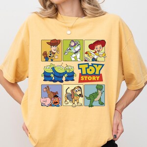May include: Yellow t-shirt featuring a colorful graphic of characters from the animated film "Toy Story." The design includes images of Woody, Buzz Lightyear, Jessie, the Aliens, and more, with the "Toy Story" logo.