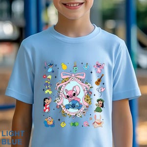May include: A light blue t-shirt with a vibrant graphic of cartoon characters, including a pink and blue alien pair in a floral frame. Other characters and objects, such as a ukulele and pineapple, are also featured. The text "LIGHT BLUE" is at the bottom.