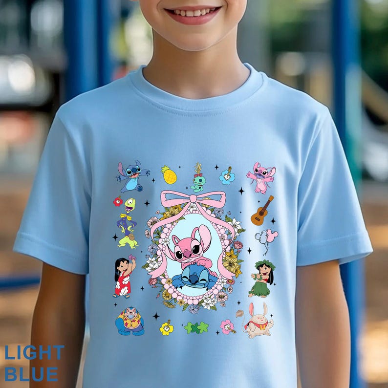 May include: A light blue t-shirt with a vibrant graphic of cartoon characters, including a pink and blue alien pair in a floral frame. Other characters and objects, such as a ukulele and pineapple, are also featured. The text "LIGHT BLUE" is at the bottom.