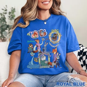 May include: Royal blue t-shirt with a vibrant graphic of Zootopia characters. The design incorporates framed portraits and the film's title. The shirt is a relaxed, casual style, suitable for everyday wear.