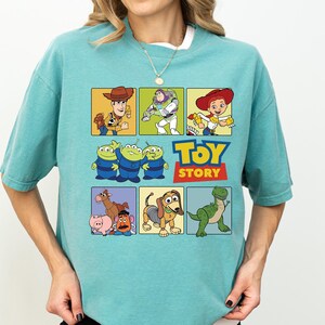 May include: A light teal t-shirt featuring a colorful graphic of Toy Story characters in square panels. The Toy Story logo is in the center. The shirt has short sleeves and a crew neck.