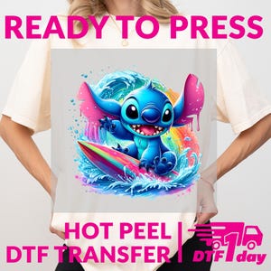 May include: A light-colored t-shirt featuring a vibrant graphic of a blue cartoon character surfing on a rainbow-colored surfboard, with a wave and rainbow background. The text "READY TO PRESS" and "HOT PEEL DTF TRANSFER" are displayed above and below the graphic.