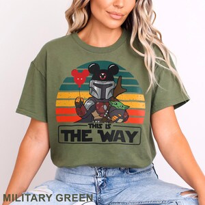 May include: Military green t-shirt featuring a graphic of The Mandalorian with Mickey Mouse ears, holding a red heart-shaped balloon and Grogu. The text "THIS IS THE WAY" is printed below the graphic. The shirt has a retro sunset design.