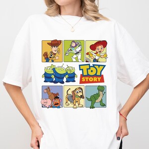 May include: White t-shirt featuring a colorful graphic of characters from the animated film "Toy Story." The design includes Woody, Buzz Lightyear, Jessie, the Aliens, the Toy Story logo, Mr. Potato Head, Slinky Dog, and Rex.