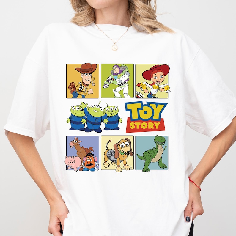 May include: White t-shirt featuring a colorful graphic of characters from the animated film "Toy Story." The design includes Woody, Buzz Lightyear, Jessie, the Aliens, the Toy Story logo, Mr. Potato Head, Slinky Dog, and Rex.