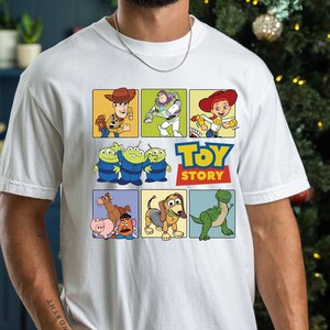 May include: White t-shirt featuring a colorful graphic of characters from the animated film "Toy Story." The design includes images of Woody, Buzz Lightyear, Jessie, the Aliens, and more, with the "Toy Story" logo in blue and yellow.