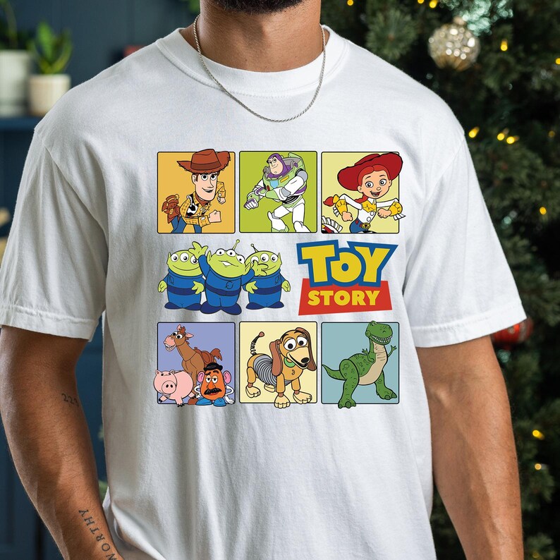 May include: White t-shirt featuring a colorful graphic of characters from the animated film "Toy Story." The design includes images of Woody, Buzz Lightyear, Jessie, the Aliens, and more, with the "Toy Story" logo in blue and yellow.