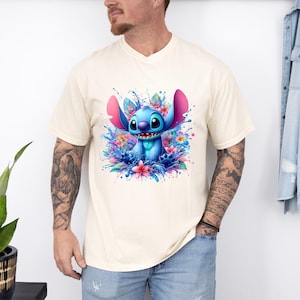 May include: Cream-colored t-shirt featuring a vibrant illustration of Stitch, surrounded by colorful flowers and water splashes. The blue alien character has large ears and a friendly expression. The design is a playful and whimsical graphic.