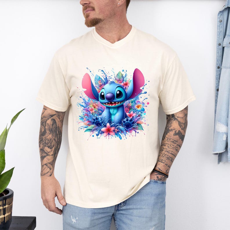 May include: Cream-colored t-shirt featuring a vibrant illustration of Stitch, surrounded by colorful flowers and water splashes. The blue alien character has large ears and a friendly expression. The design is a playful and whimsical graphic.