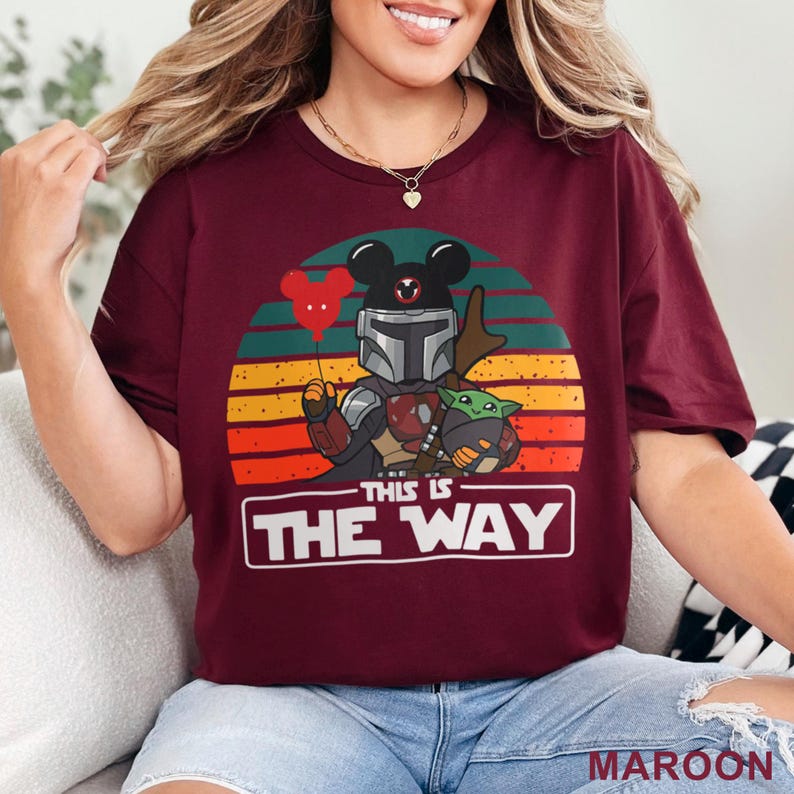 May include: Maroon t-shirt featuring a graphic of a Mandalorian character with Mickey Mouse ears holding a red balloon and Baby Yoda. The text "THIS IS THE WAY" is displayed in a white rectangle. The background has a sunset design.