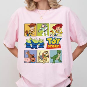 May include: A light pink t-shirt featuring a colorful graphic of characters from the animated film "Toy Story." The design includes images of Woody, Buzz Lightyear, Jessie, the Aliens, and others, with the "Toy Story" logo.