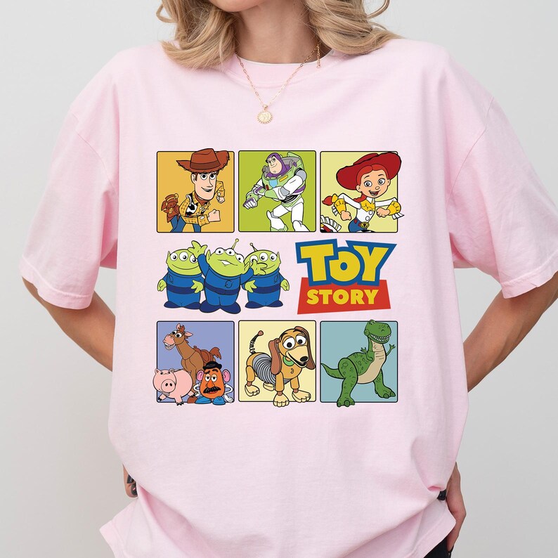 May include: A light pink t-shirt featuring a colorful graphic of characters from the animated film "Toy Story." The design includes images of Woody, Buzz Lightyear, Jessie, the Aliens, and others, with the "Toy Story" logo.