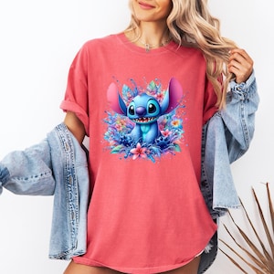 May include: A coral-colored t-shirt featuring a vibrant illustration of Stitch from Lilo & Stitch. The blue alien is surrounded by colorful flowers and water splashes. The shirt has a crew neck and short sleeves.