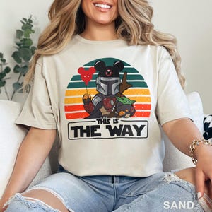 May include: Sand-colored t-shirt featuring a graphic of The Mandalorian with a Mickey Mouse-eared helmet holding a red heart balloon and Grogu. The design is set against a sunset backdrop with the text "THIS IS THE WAY".