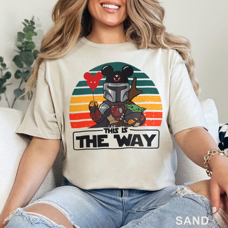 May include: Sand-colored t-shirt featuring a graphic of The Mandalorian with a Mickey Mouse-eared helmet holding a red heart balloon and Grogu. The design is set against a sunset backdrop with the text "THIS IS THE WAY".