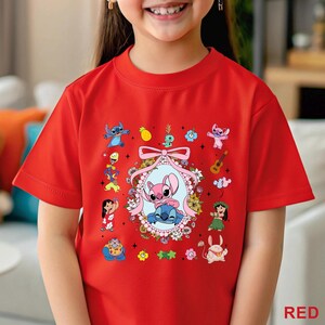 May include: A red t-shirt with a vibrant cartoon graphic. The design showcases a central oval frame with two characters, encircled by flowers, a bow, and other characters. The word "RED" is printed in the bottom right corner of the t-shirt.