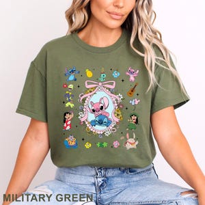 May include: Military green t-shirt with a vibrant graphic of animated characters. The design showcases a central oval frame with two characters, surrounded by other characters and floral details. The text "MILITARY GREEN" is displayed at the bottom of the shirt.