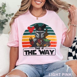 May include: Light pink t-shirt featuring a graphic of a Mandalorian character with a Mickey Mouse head, holding a red heart balloon, and Grogu. The design is set against a sunset background with the text "THIS IS THE WAY".