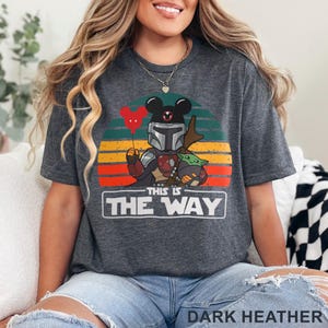 May include: Dark heather gray t-shirt featuring a graphic of a Mandalorian character with a Mickey Mouse head, holding a red heart balloon, and Grogu. The text "THIS IS THE WAY" is displayed below the graphic, with a sunset background.