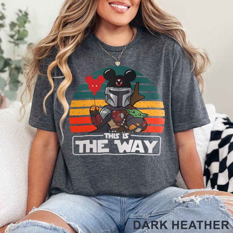 May include: Dark heather gray t-shirt featuring a graphic of a Mandalorian character with a Mickey Mouse head, holding a red heart balloon, and Grogu. The text "THIS IS THE WAY" is displayed below the graphic, with a sunset background.