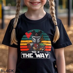 May include: Black t-shirt featuring a graphic of a Mandalorian character with a Mickey Mouse head, holding a red heart balloon, and Grogu. The design is set against a sunset-colored background with the text "THIS IS THE WAY" in a white rectangle.