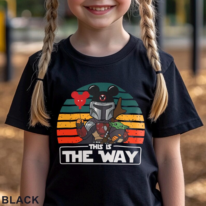 May include: Black t-shirt featuring a graphic of a Mandalorian character with a Mickey Mouse head, holding a red heart balloon, and Grogu. The design is set against a sunset-colored background with the text "THIS IS THE WAY" in a white rectangle.