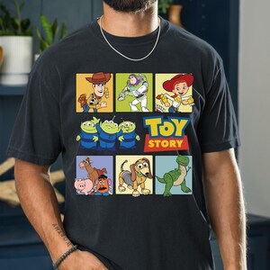 May include: Black t-shirt featuring a colorful graphic of characters from the animated film "Toy Story." The design includes Woody, Buzz Lightyear, Jessie, the Aliens, Hamm, Slinky Dog, and Rex, with the "Toy Story" logo.